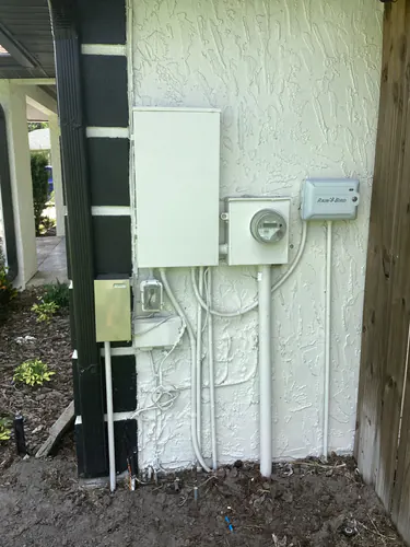 Outdoor service wall of a Longwood home with the meter socket, main panel, irrigation controller and PVC risers into the ground