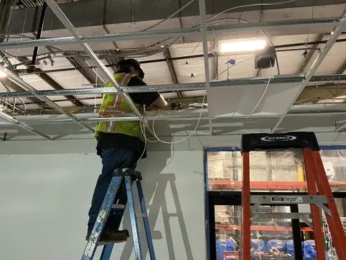 Electrician on a ladder installing boxes above the ceiling grid in the EMS warehouse office area