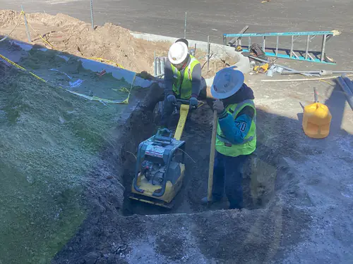 Two workers compacting backfill in a trench with a plate compactor at Massey Cadillac