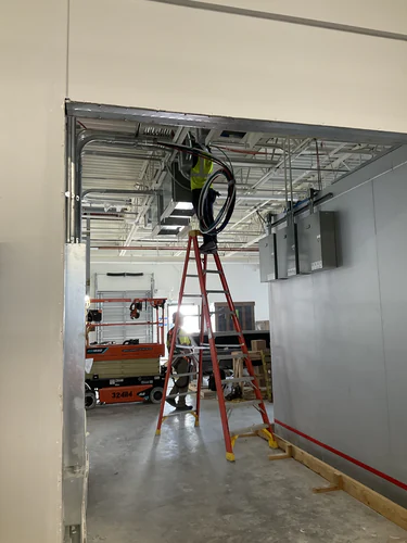 Electrician on a ladder pulling MC cable above the ceiling beside the panels at Lady Lake Market